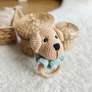 以下が含まれることがあります： 青い編み物の襟が付いた、茶色の編み物の犬のガラガラのおもちゃ。ガラガラは白いふわふわの表面の上にあります。