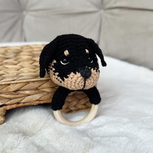 May include: A handcrafted crochet dog rattle in black and beige. The dog's head has black ears and a beige muzzle. The rattle includes a wooden ring for easy handling. The toy is on a woven basket and a white, fluffy surface.