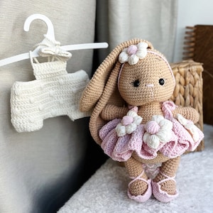 Puede incluir: Una muñeca de conejo de crochet que lleva un vestido rosa con flores blancas. El conejo tiene pelo marrón y ojos negros. La muñeca está sentada sobre una superficie blanca y esponjosa.