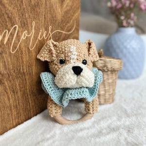 Puede incluir: Un sonajero de crochet de cachorro marrón y blanco con un collar de crochet azul claro. El sonajero tiene un anillo de madera.