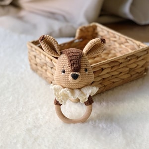 Op de afbeelding: Een gehaakte hertenrammelaar met een houten ring. Het hert is bruin en beige met een witte gehaakte kraag.