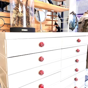 Könnte beinhalten: Weiße Kommode mit acht Schubladen und roten, pilzförmigen Knöpfen. Jeder Knopf hat weiße Punkte.