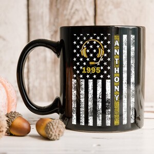 May include: Black ceramic coffee mug with a white and black American flag design. The flag has a yellow stripe with the name "ANTHONY" and the year "1995" in gold lettering. A gold headphone icon is on the flag.