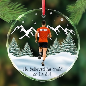 May include: A clear, round Christmas ornament with a red ribbon. The ornament features a runner in an orange shirt with the name "ANTONY" on the back, running in a snowy mountain scene. The text "He believed he could so he did" is at the bottom.