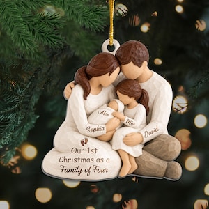 May include: A carved-style hanging ornament depicting a family of four with brown hair, wearing cream-colored clothing. The figures are labeled with the names Sophia, Alex, Mia, and David. It features the text Our 1st Christmas as a Family of Four.