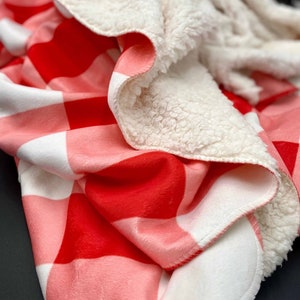 May include: A red and white checked throw blanket with a soft, white sherpa lining. The blanket is folded in half, showing both sides.