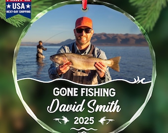 May include: A clear, round glass ornament with a photo of a man holding a fish. The ornament has the text "GONE FISHING David Smith 2025" and a red ribbon for hanging. The ornament also has the text "PRINT IN USA NEXT-DAY SHIPPING".