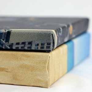 May include: Two canvases stacked on top of each other. The top canvas has a dark blue and gray patterned design. The bottom canvas is a light beige color.