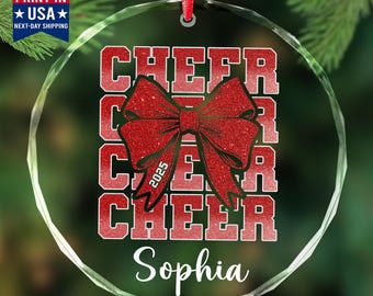 May include: A clear glass ornament with the word "CHEER" in red block letters, a red glitter bow, and the name "Sophia" in white script. The year "2025" is on the bow. A red ribbon is attached for hanging.