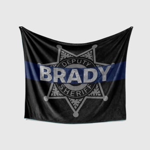 May include: Black fleece blanket with a blue and white sheriff's star badge. The badge has the text "DEPUTY SHERIFF" and the name "BRADY" in the center.