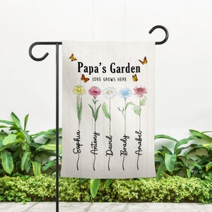 May include: A garden flag with the text "Papa's Garden Love Grows Here" and a drawing of six flowers with names written below each flower: Sophia, Antony, David, Braidy, Amabel.  The flag is beige with a white background and has two orange butterflies flying above the text.