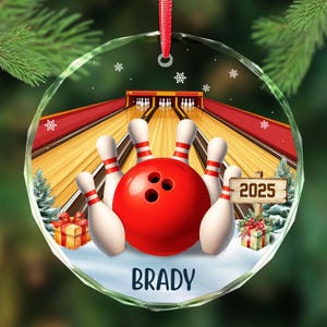 May include: A round, clear glass Christmas ornament featuring a bowling scene. A red bowling ball is surrounded by white and red striped pins. The word "BRADY" is at the bottom, and the year "2025" is on a sign.