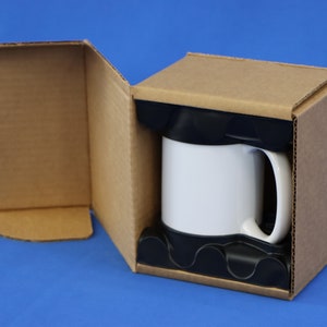 May include: A white coffee mug with a black handle is nestled in a cardboard box with protective black foam inserts. The box is open, revealing the mug against a blue background. The box is made of corrugated cardboard.