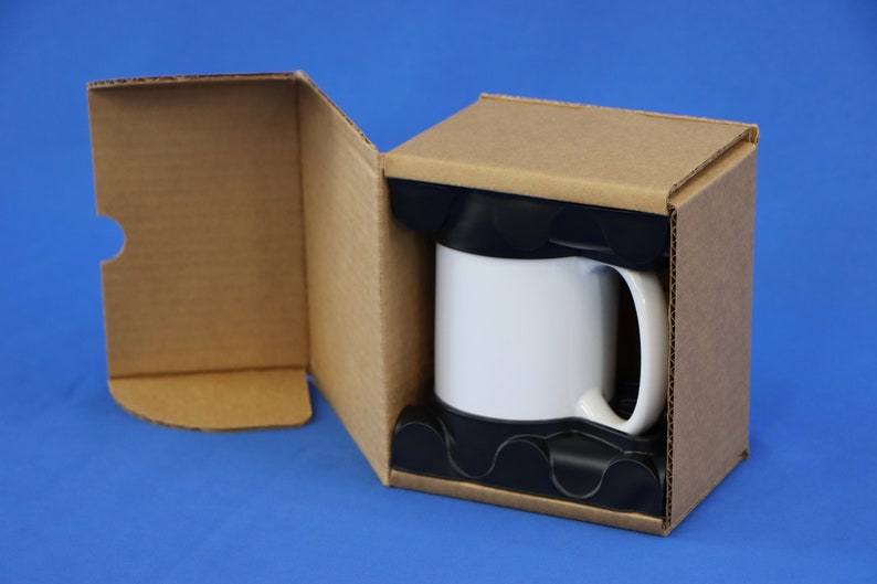 May include: A white coffee mug with a black handle is nestled in a cardboard box with protective black foam inserts. The box is open, revealing the mug against a blue background. The box is made of corrugated cardboard.