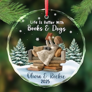 May include: A clear, round Christmas ornament with a red ribbon. The ornament features a woman reading on a couch with a dog, surrounded by pine trees and snowflakes. Text reads "Life Is Better With Books & Dogs", "Olivia & Rockie 2025".