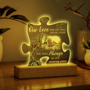 May include: A puzzle piece-shaped acrylic light display on a rectangular wooden base. It features a printed photograph of a person, a tree with red birds, and the text: Our Love FOR YOU WILL NEVER CEASE YOU WILL Always BE OUR missing piece.