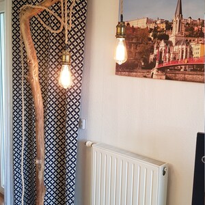 May include: A rustic floor lamp made from a natural branch with three hanging light bulbs. The branch is secured to the floor with a rope and the light bulbs are suspended from the branch with rope. The lamp is in a room with a white radiator and a black and white patterned curtain.
