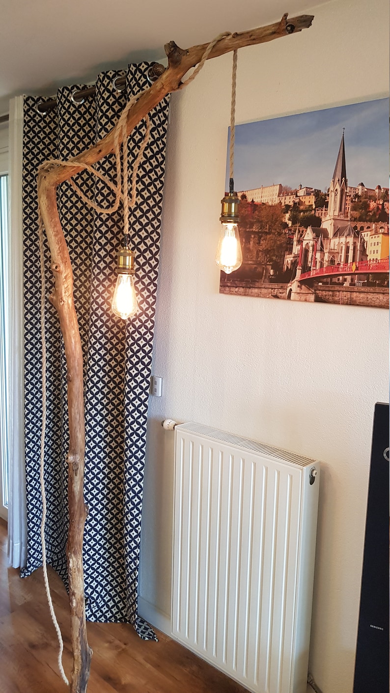 May include: A rustic floor lamp made from a natural branch with three hanging light bulbs. The branch is secured to the floor with a rope and the light bulbs are suspended from the branch with rope. The lamp is in a room with a white radiator and a black and white patterned curtain.