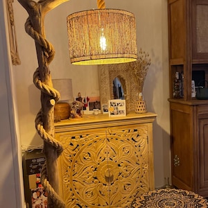 May include: A wooden floor lamp with a rope wrapped around the base and a woven shade. The lamp is next to a carved wooden cabinet with two doors and a woven footstool with a black and white pattern.