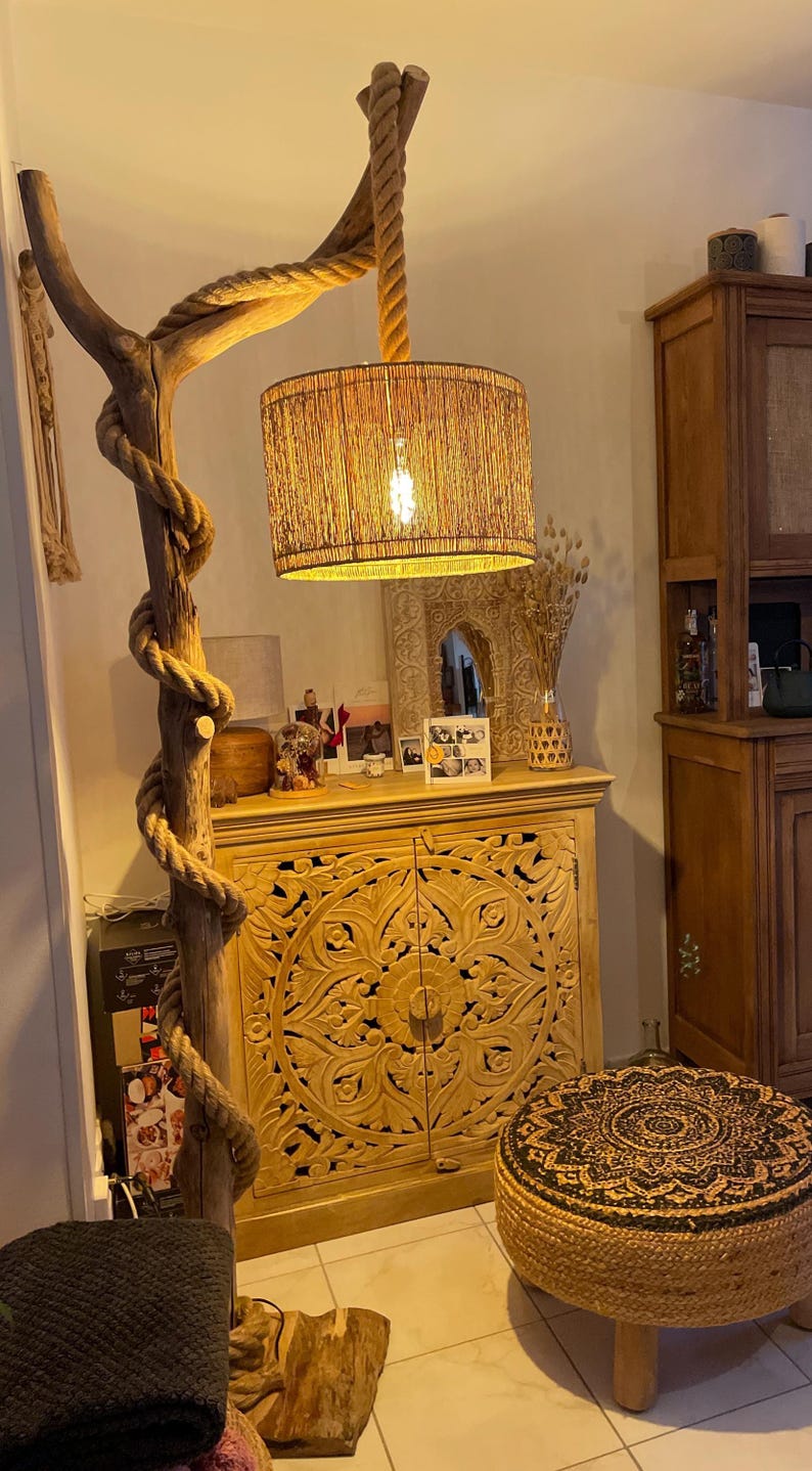 May include: A wooden floor lamp with a rope wrapped around the base and a woven shade. The lamp is next to a carved wooden cabinet with two doors and a woven footstool with a black and white pattern.