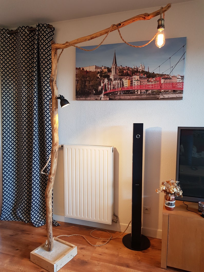May include: A rustic floor lamp made from a natural branch with two light fixtures. The lamp has a rope cord and a wooden base. The lamp is lit and casting a warm glow on the surrounding room.