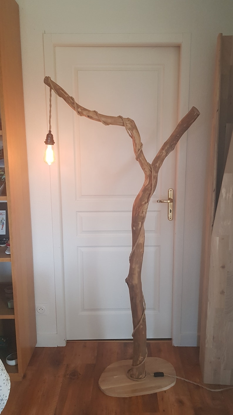 May include: A rustic floor lamp made from a natural branch with a light bulb hanging from a cord. The lamp is on a wooden base and has a natural rope wrapped around the branch.