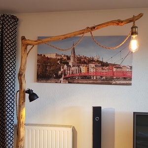May include: A rustic wooden floor lamp with a natural branch design. The lamp has two light fixtures, one with a black shade and the other with a clear glass bulb. The lamp is positioned in front of a canvas print of a city skyline with a red bridge.