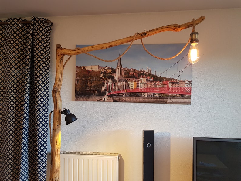 May include: A rustic wooden floor lamp with a natural branch design. The lamp has two light fixtures, one with a black shade and the other with a clear glass bulb. The lamp is positioned in front of a canvas print of a city skyline with a red bridge.