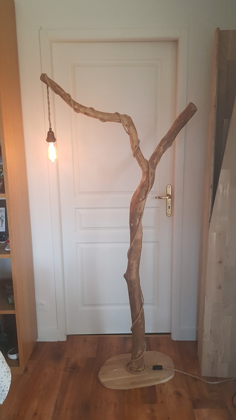 May include: A wooden floor lamp shaped like a tree branch with a light bulb hanging from a cord. The lamp is in a room with a white door and wooden floor.