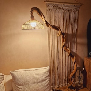 May include: A floor lamp with a woven shade and a natural wood base shaped like a tree branch. The lamp is lit and casting a warm glow. The lamp is in front of a macrame wall hanging.