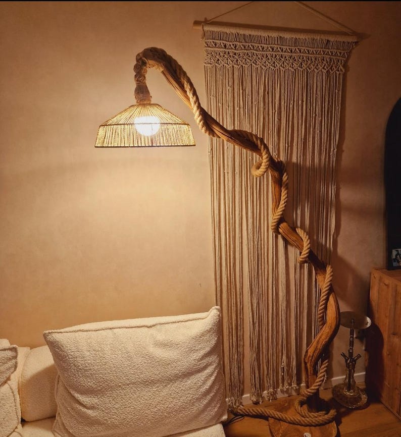 May include: A floor lamp with a woven shade and a natural wood base shaped like a tree branch. The lamp is lit and casting a warm glow. The lamp is in front of a macrame wall hanging.