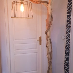 May include: A rustic floor lamp made from a natural branch wrapped in rope with a wire cage lampshade. The lamp is lit with a light bulb and is standing on a wooden base.