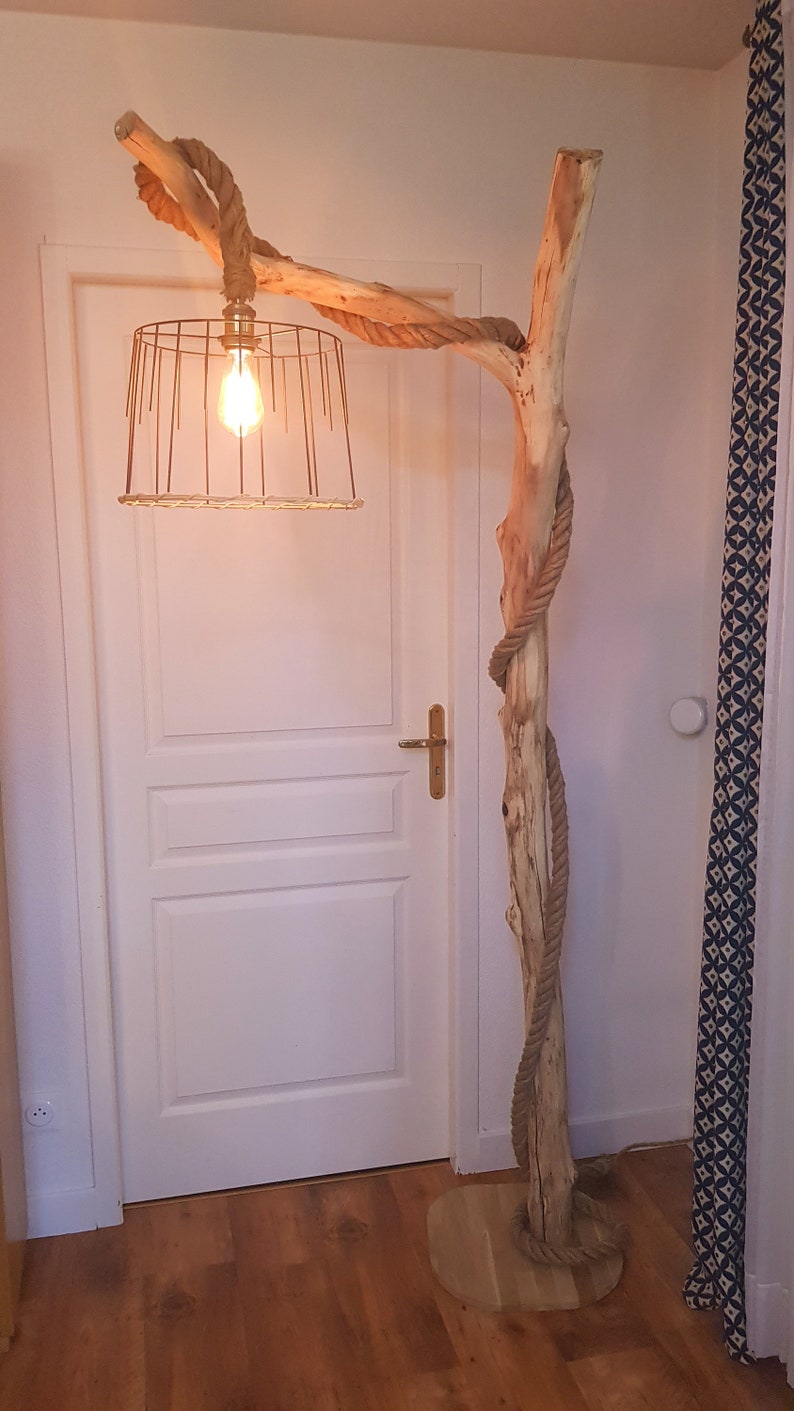 May include: A rustic floor lamp made from a natural branch wrapped in rope with a wire cage lampshade. The lamp is lit with a light bulb and is standing on a wooden base.