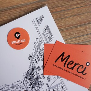 May include: Two orange and black business cards with the text "Merci de me soutenir dans cette nouvelle aventure!" and a small black and white logo of a location pin with a circle around it. The cards are on top of a white sheet of paper with a black and white drawing of a building.