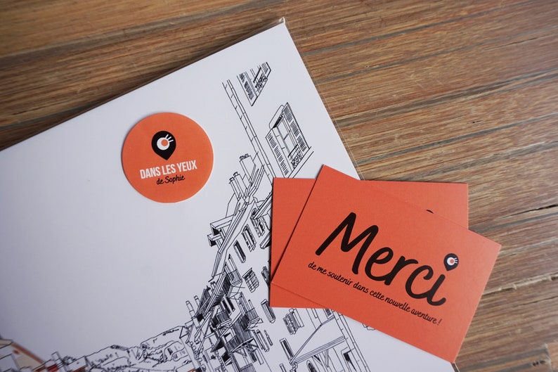 May include: Two orange and black business cards with the text "Merci de me soutenir dans cette nouvelle aventure!" and a small black and white logo of a location pin with a circle around it. The cards are on top of a white sheet of paper with a black and white drawing of a building.