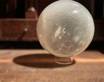 Puede incluir: Un objeto esférico translúcido con una superficie texturizada, posiblemente una esfera de vidrio decorativa o una bola de cristal. Se apoya sobre una pequeña base transparente, proyectando una sombra sobre la superficie de madera. La estética general es de elegancia y misterio.