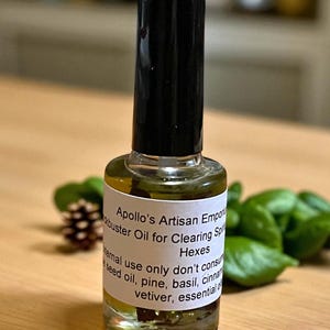 May include: A clear glass bottle with a black cap, filled with a yellow liquid. The label reads "Apollo's Artisan Emporium" and "Hexes." The bottle is surrounded by green leaves and a pine cone.
