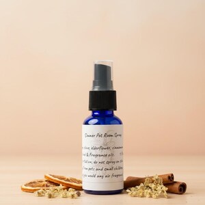 Op de afbeelding: Een blauwe glazen fles Simmer Pot Room Spray met een zwarte sproeikop. Het etiket vermeldt de ingrediënten. Gedroogde sinaasappelschijfjes, kaneelstokjes en gedroogde bloemen zijn rond de fles gerangschikt.