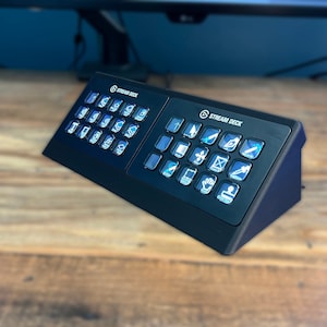 May include: A black Stream Deck with two rows of buttons, each with a different icon. The buttons are illuminated with a blue light. The Stream Deck is sitting on a wooden surface.