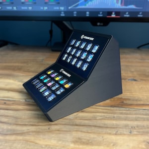 May include: A black Stream Deck device with multiple illuminated buttons, arranged in two tiers. The device is angled, with the top section slightly raised. The Stream Deck logo is visible. The device sits on a wooden surface.