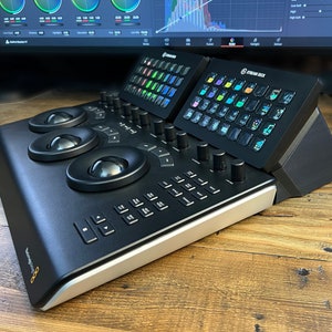 May include: A black professional audio mixing console with three trackballs, a row of knobs, and a keypad. Two black Stream Deck controllers with colour-coded buttons are attached to the console.