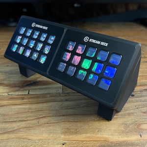 Op de afbeelding: Een zwarte Stream Deck met twee rijen aanpasbare knoppen. Elke knop heeft een klein LCD-scherm dat verschillende pictogrammen en afbeeldingen weergeeft. De Stream Deck staat op een houten oppervlak.