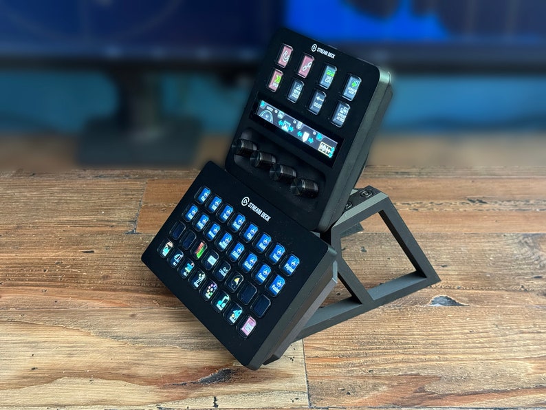 May include: A black Stream Deck XL with a smaller Stream Deck Mini stacked on top. Both devices have a variety of buttons with icons and text labels. The devices are on a wooden surface.