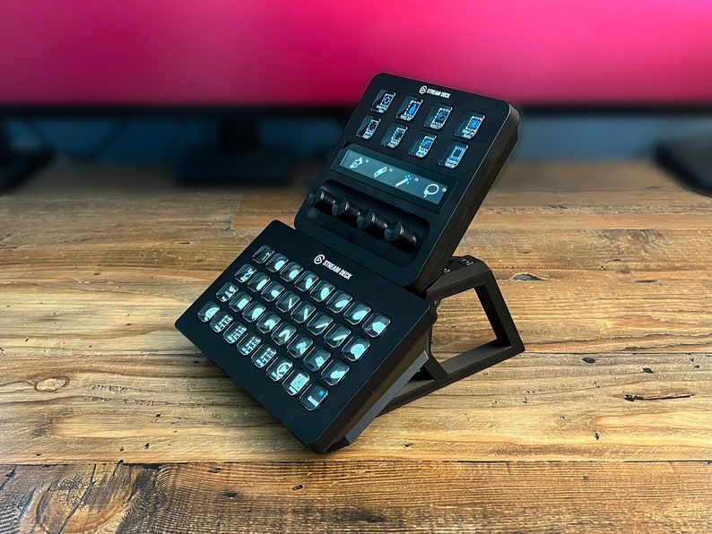 May include: A black Stream Deck with a stand, featuring a grid of customizable buttons with icons and a small LCD screen with a few buttons and knobs.