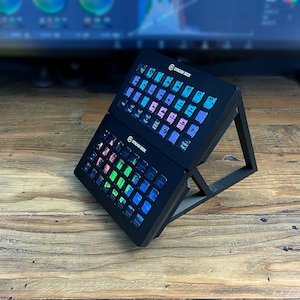 May include: Two black Stream Deck devices with colorful buttons are stacked on a black stand. The stand is on a wooden surface.