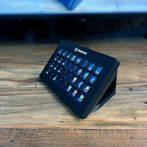 May include: A black Stream Deck with a stand, featuring a grid of customisable buttons with icons for various streaming applications.
