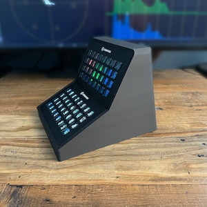 May include: A grey Stream Deck device with numerous illuminated buttons. The device is angled, with a black top section displaying the buttons. The Stream Deck logo is visible. It rests on a wooden surface.