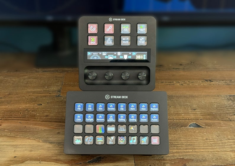 May include: Two black Stream Deck devices with a variety of buttons and a small LCD screen on the top device. The buttons have various icons and text labels, including "File", "Edit", "Trim", "Timeline", "Clip", "Mark", "View", "Playback", "Color", "Brightness", "Microphone", "Mute", "Editing", "Grading", "Sound", "Fusion", "Launch", "Undo", "Unloaded", "Gallery", "Scene", "Camera", "Mic", "Music", "Browser", "Game", "Social", "Timer", "Text", "Scene", "Source", "Settings", "Profile", "Stream Deck".