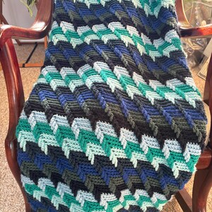 May include: A handmade crochet blanket in shades of blue, teal, and gray, draped over a wooden chair. The blanket features a textured, chevron-like pattern. The color palette creates a cozy and inviting feel.