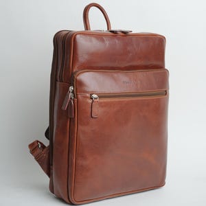 Puede incluir: Una mochila de cuero marrón con un bolsillo frontal con cremallera y el nombre de la marca "BRIGHTON" grabado en la parte delantera.